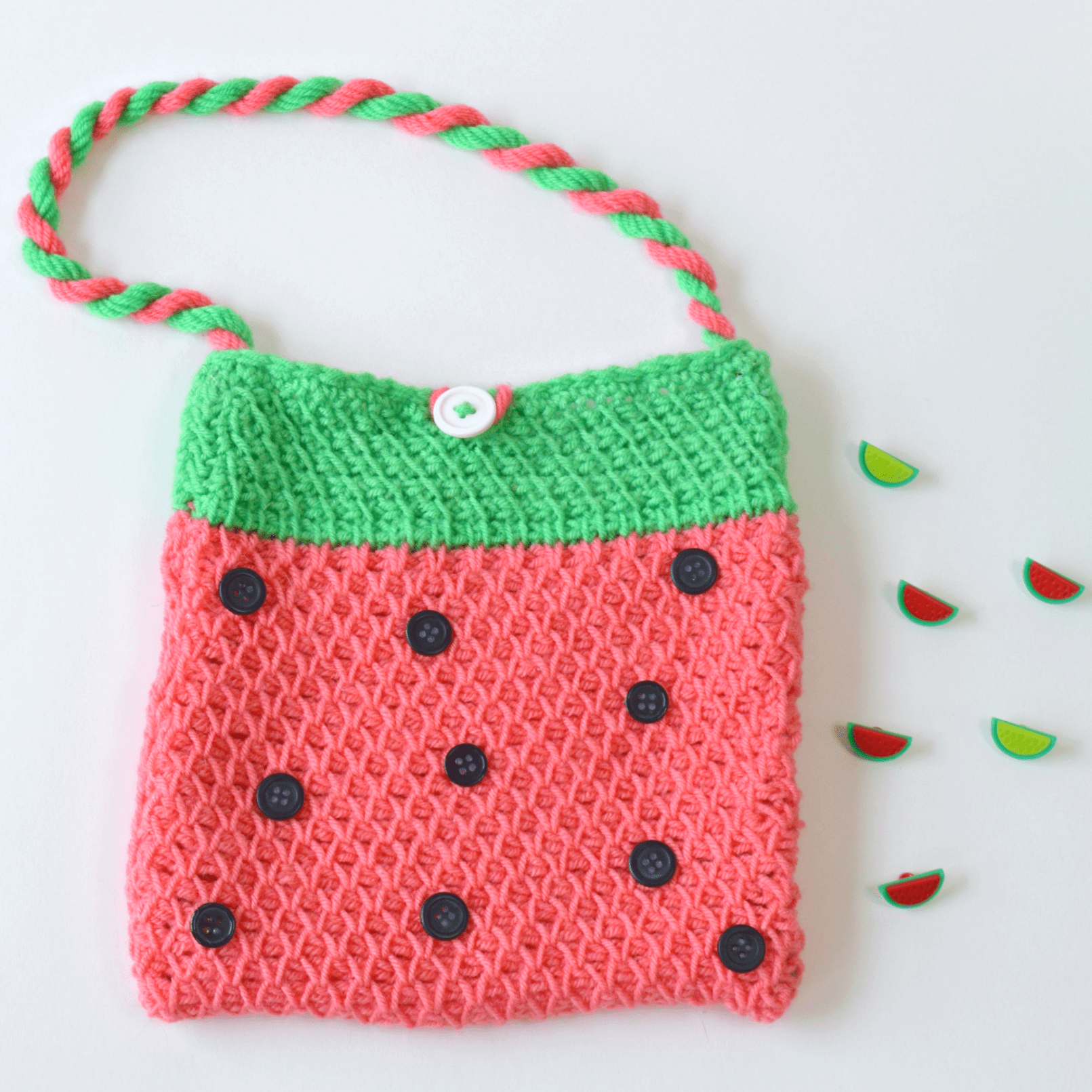 Colorful Tunisian watermelon bag made with Tunisian simple stitch featuring a pink body with black button 'seeds,' green top edge, and a twisted pink and green yarn strap. Fun and fruity crochet accessory perfect for summer or kids' bags.