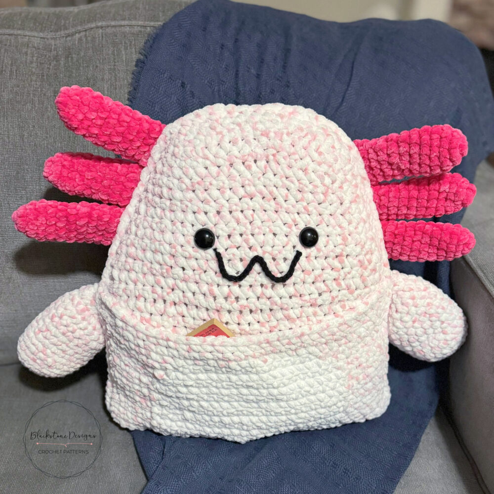 Crochet Axolotl Pillow with Book Pocket belly, made with light pink yarn for the body and dark pink for the external gills
