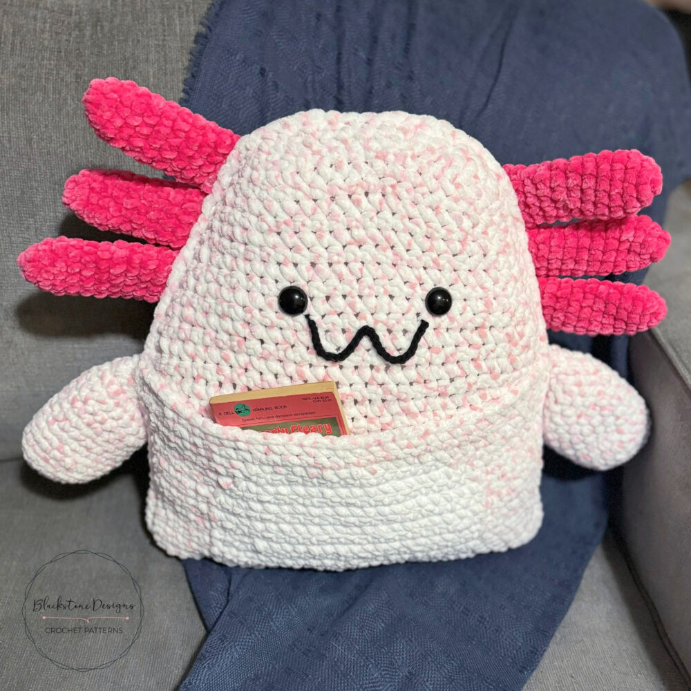 Crochet Axolotl Pillow with Book Pocket belly, made with light pink yarn for the body and dark pink for the external gills