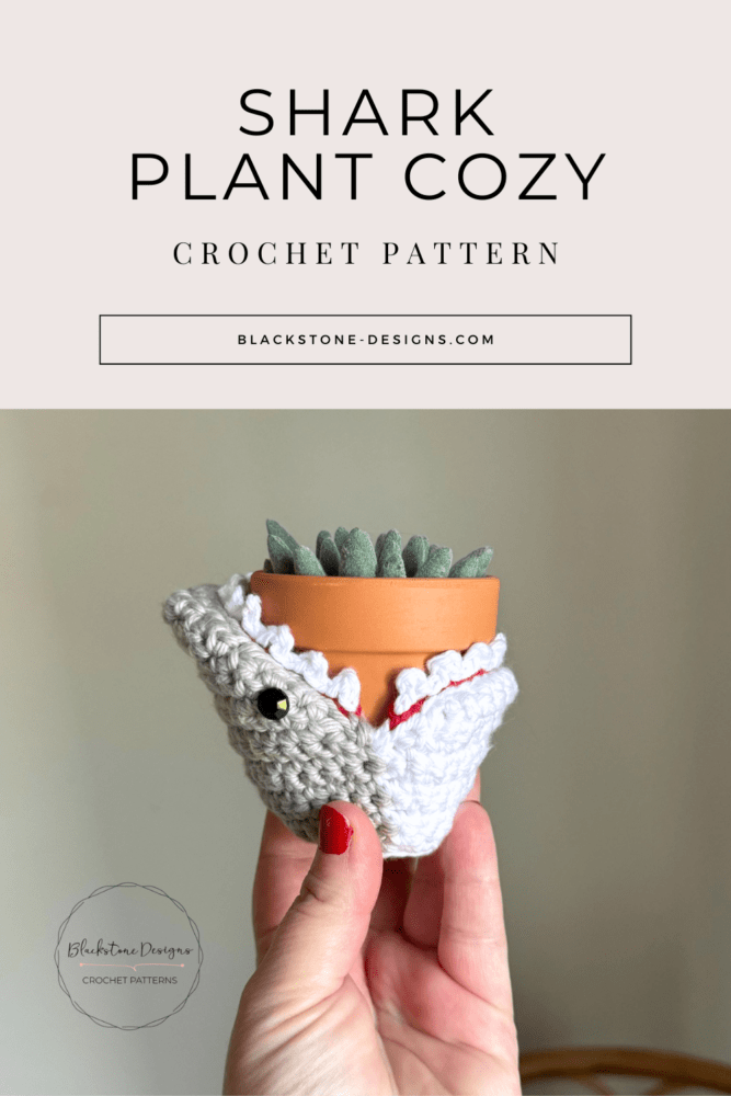 shark crochet plant cozy wrapped around a mini terracotta pot with succulent inside, featuring gray yarn, white teeth trim, and black safety eyes.
