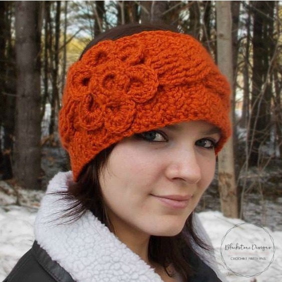 Woman wearing orange ear warmer featuring braided cables and a big flower with a snowy background