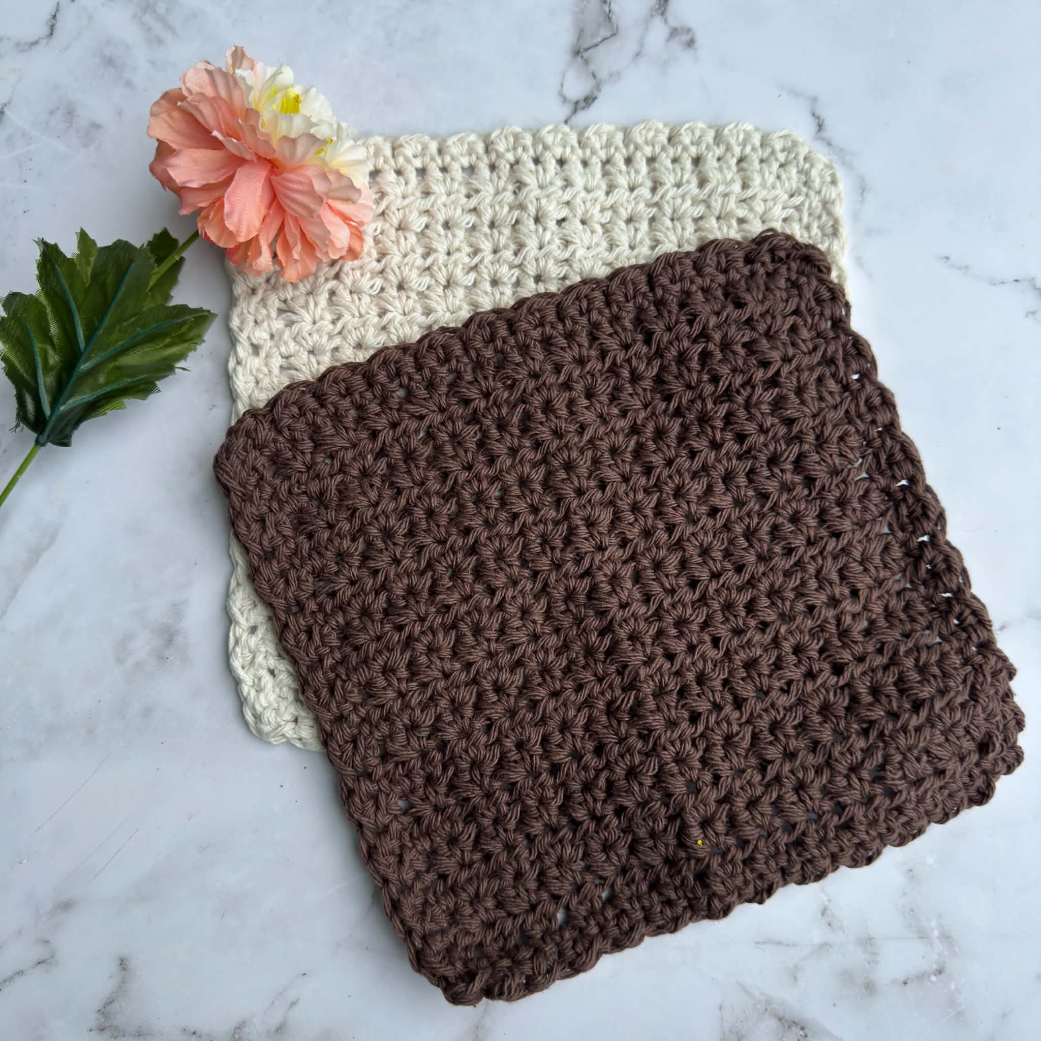Cream and brown Magnolia Heirloom crochet dishcloths stacked on a white marble background with a peach magnolia flower.