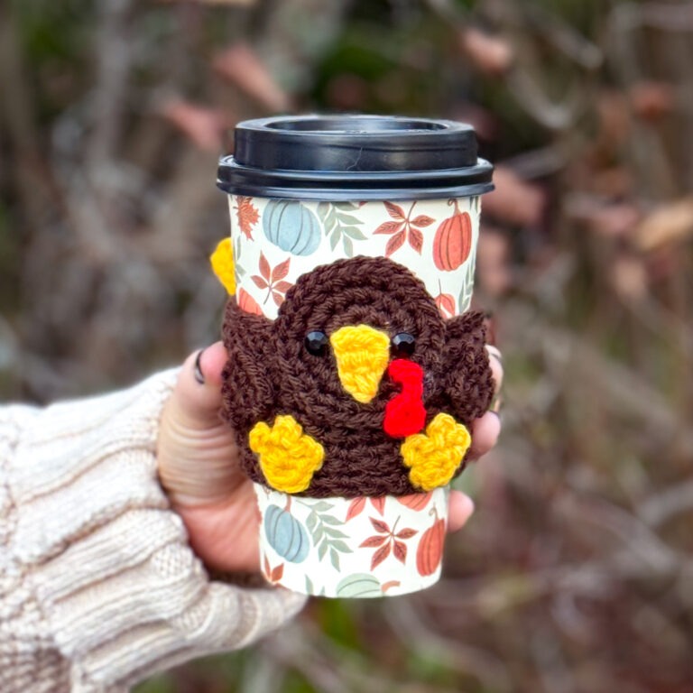Handmade crochet turkey coffee sleeve with brown body, yellow beak and feet, red wattle, and black eyes wrapped around a fall-themed takeaway coffee cup.